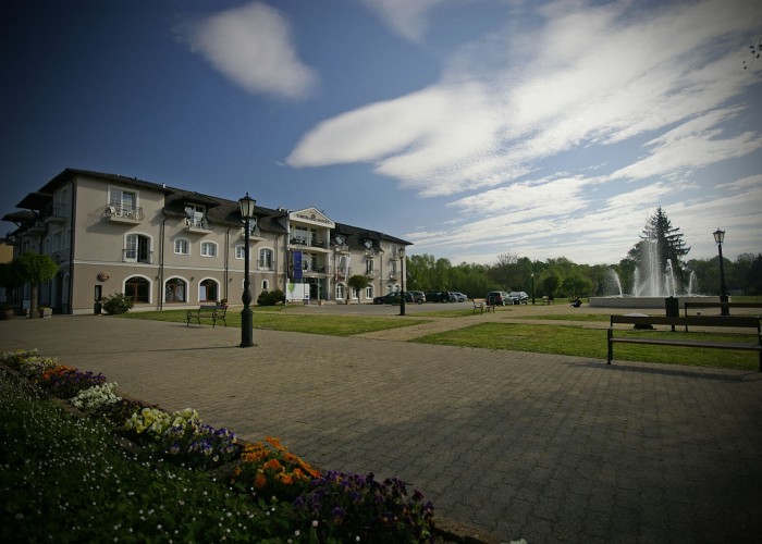 A Harkányi Xavin szállodájának utcaképe, háttérben a városi parkkal és szökőkúttal, valamint parkolóval.